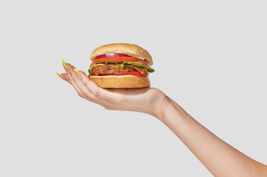 A white woman's hand with long acrylic nails holds a plant-based sandwich. 